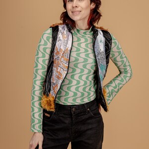 May include: A woman wearing a black puffer vest with a colorful patterned lining, a green and white patterned long-sleeved shirt, and black jeans.