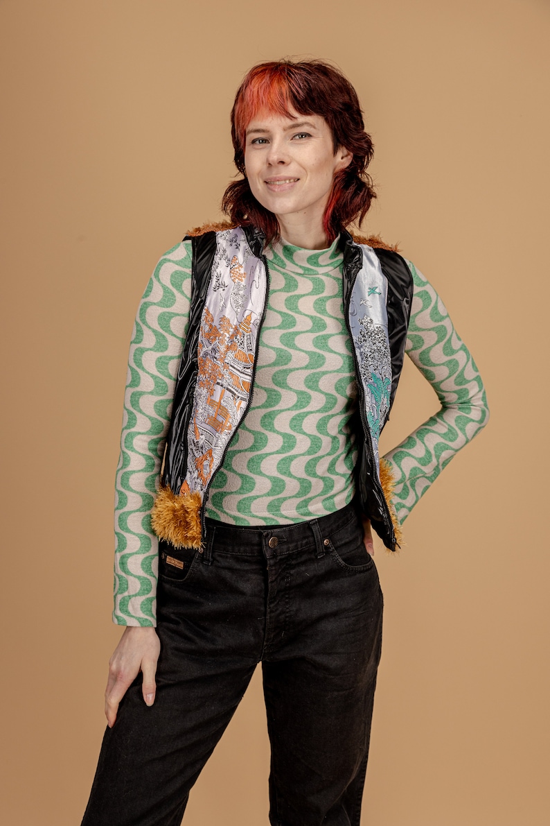 May include: A woman wearing a black puffer vest with a colorful patterned lining, a green and white patterned long-sleeved shirt, and black jeans.