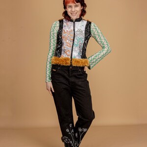 May include: A person wearing a black and green patterned vest with a brown fur trim, black jeans with white floral embroidery, and brown boots.