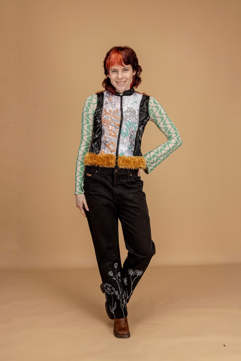 May include: A person wearing a black and green patterned vest with a brown fur trim, black jeans with white floral embroidery, and brown boots.