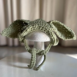 May include: A light green crochet hat with large, floppy ears and tie strings. The hat is displayed on a white mushroom-shaped stand. The hat's texture is visible, showcasing the handmade quality of the item.