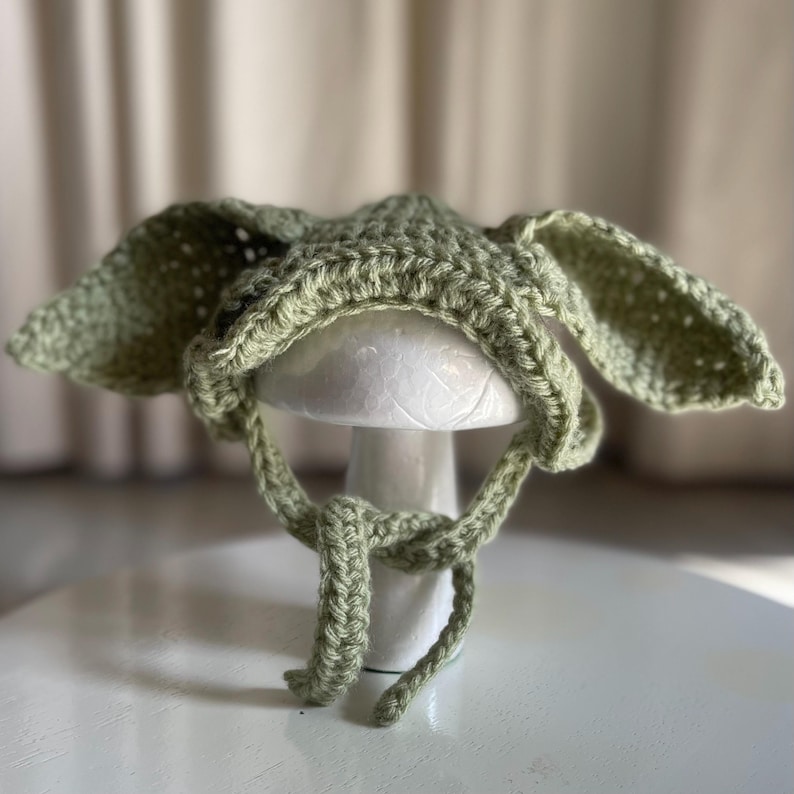 May include: A light green crochet hat with large, floppy ears and tie strings. The hat is displayed on a white mushroom-shaped stand. The hat's texture is visible, showcasing the handmade quality of the item.