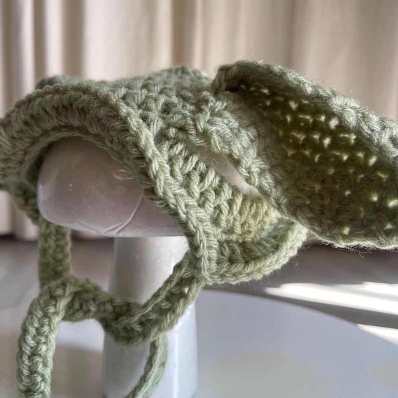 May include: A light green, crocheted hat with long, floppy ears. The hat is made of a chunky yarn and has ties that hang down. The hat is displayed on a white mannequin head.