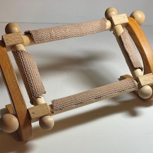 May include: A wooden embroidery hoop with a rectangular frame. The frame is made of light brown wood with rounded wooden knobs at each corner. Beige fabric is stretched across the frame, providing a surface for needlework. The hoop is designed to hold fabric taut.