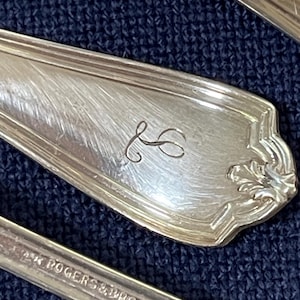 May include: Close-up of antique silverware with a decorative floral design and an engraved initial. The spoon and fork are silver-toned and rest on a dark blue textured surface. The silverware is marked "WM ROGERS & BRO. A1".