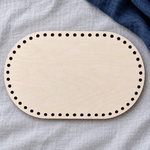May include: An oval-shaped light-colored wooden base featuring a smooth, flat surface and a series of evenly spaced, circular holes punched along the entire perimeter, designed for crafting or basket weaving projects.