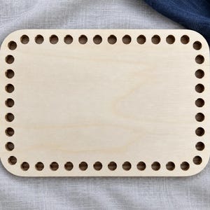 May include: A rectangular light-colored wooden base with rounded corners, featuring a series of evenly spaced circular holes punched along the entire perimeter. The surface of the wood is smooth and displays a natural, light beige grain pattern.