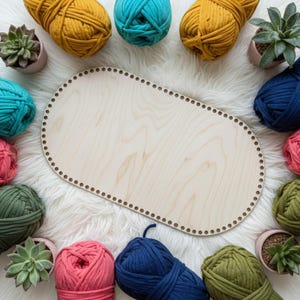 May include: An oval-shaped wooden base with a series of small, evenly spaced holes along the perimeter, surrounded by multiple balls of yarn in shades of mustard yellow, teal, navy blue, olive green, and pink, along with small potted succulents.