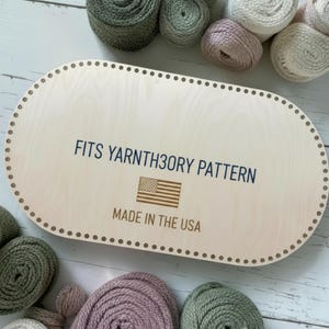 May include: An oval-shaped light wood base with a series of small holes along the perimeter. The center features the text FITS YARNTH3ORY PATTERN, a small American flag graphic, and the text MADE IN THE USA printed in dark blue and brown ink.