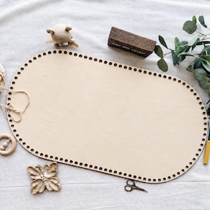 May include: A large, oval-shaped light-colored wooden base featuring a series of evenly spaced holes along the entire perimeter, designed for crafting projects. The surface is smooth and flat, showcasing the natural grain of the wood material.
