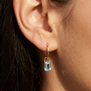 May include: A gold hoop earring with a rectangular, light blue gemstone pendant. The earring is attached to an ear. The gemstone is a light blue color and is set in a gold frame. The earring is a simple, elegant design.