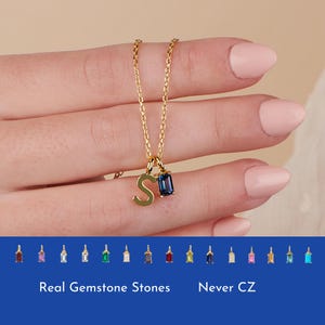 May include: Gold necklace with a letter 'S' charm and a blue gemstone pendant. The necklace is shown on a hand with a light pink nail polish. The image also shows a row of different colored gemstones, labeled 'Real Gemstone Stones' and 'Never CZ'.