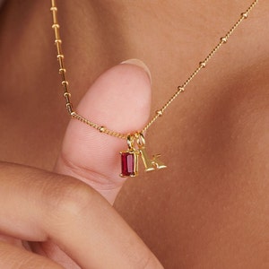 May include: A gold chain necklace with a small red gemstone pendant and a gold letter 'K' charm.
