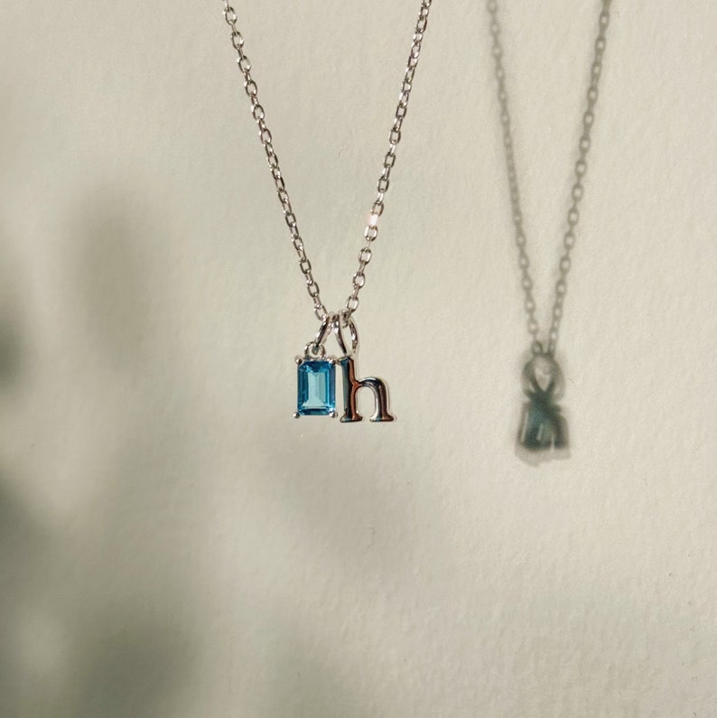 K&ouml;nnte beinhalten: Eine silberne Halskette mit einem blauen Edelstein und einem silbernen Anh&auml;nger mit dem Buchstaben 'h'.