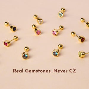 May include: Assortment of gold stud earrings with various colored gemstones. The earrings feature a small round gemstone set in a gold setting with a gold post and back. The text "Real Gemstones, Never CZ" is displayed below the earrings.