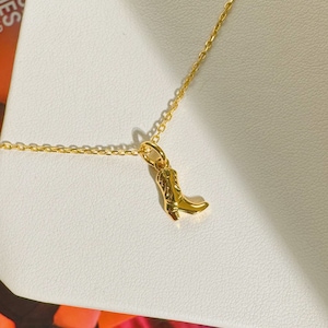 May include: A gold-colored necklace featuring a small, detailed cowboy boot pendant. The boot has intricate design elements and is suspended from a delicate chain. The jewelry is displayed on a white surface.