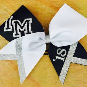 May include: Black and white cheer bow with silver glitter accents. The bow has the letters "IM" in silver glitter on the black side and the number "18" in silver glitter on the white side.