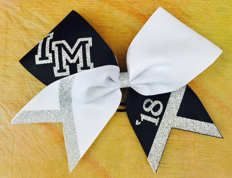 May include: Black and white cheer bow with silver glitter accents. The bow has the letters "IM" in silver glitter on the black side and the number "18" in silver glitter on the white side.