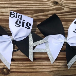 May include: Two black and white cheer bows with silver glitter accents. The larger bow has the text "Big Sis" on the white portion. The smaller bow has the text "Lil Sis" on the white portion.