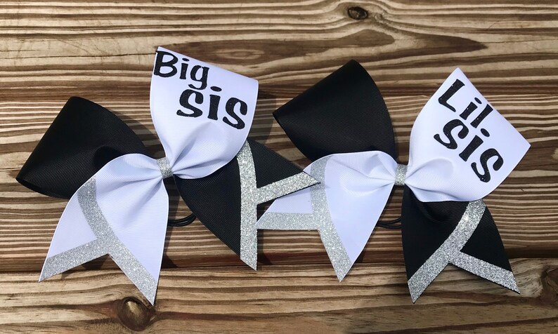 May include: Two black and white cheer bows with silver glitter accents. The larger bow has the text "Big Sis" on the white portion. The smaller bow has the text "Lil Sis" on the white portion.