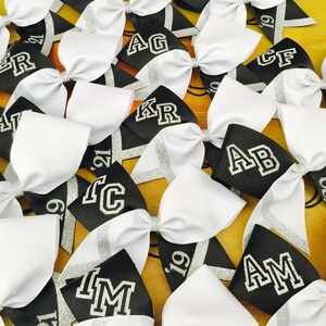 May include: A collection of black and white cheer bows with silver glitter accents. Each bow has a different letter or initials embroidered on it. The bows are arranged in a random pattern on a wooden surface.
