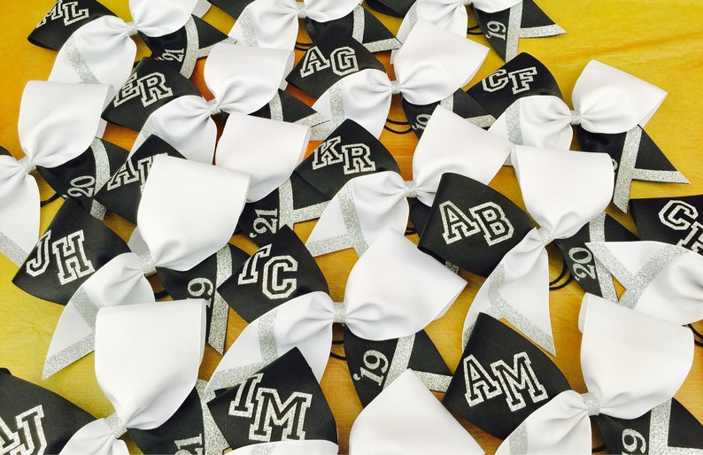 May include: A collection of black and white cheer bows with silver glitter accents. Each bow has a different letter or initials embroidered on it. The bows are arranged in a random pattern on a wooden surface.