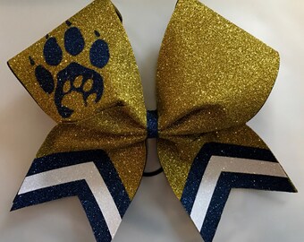 May include: A large, glittery gold cheerleading bow with a navy blue paw print and chevron design. The bow has a black elastic band and a navy blue center knot. The bow is likely used for cheerleading or dance.