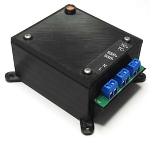 Może przedstawiać: Czarny element elektroniczny o prostokątnym kształcie i teksturowanej powierzchni. Posiada zieloną płytkę drukowaną z niebieskimi złączami i etykietami, takimi jak "TC+", "TC-", "SSR+", "SSR-", "L" i "N". Element ma cztery n&oacute;żki montażowe.