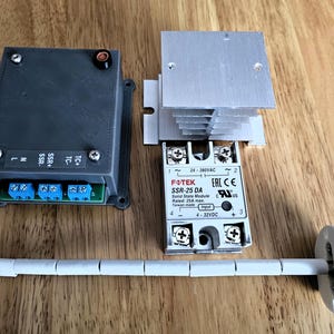 May include: A collection of electronic components, including a grey box with blue terminals, a silver heat sink, and a white ceramic tube. The components are arranged on a wooden surface. The text "FOTEK SSR-25 DA" is visible.