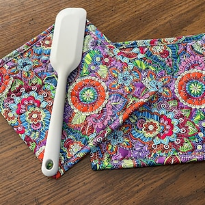 May include: A white silicone spatula rests on two colorful, patterned pot holders. The pot holders feature a vibrant floral design with orange, purple, blue, and green hues. The spatula is angled across the pot holders, suggesting kitchen use.