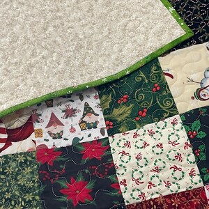 May include: A patchwork quilt with a variety of Christmas-themed patterns. The quilt features squares with gnomes, poinsettias, holly, and wreaths. The border is green, and the backing is a light beige with a snowflake pattern.