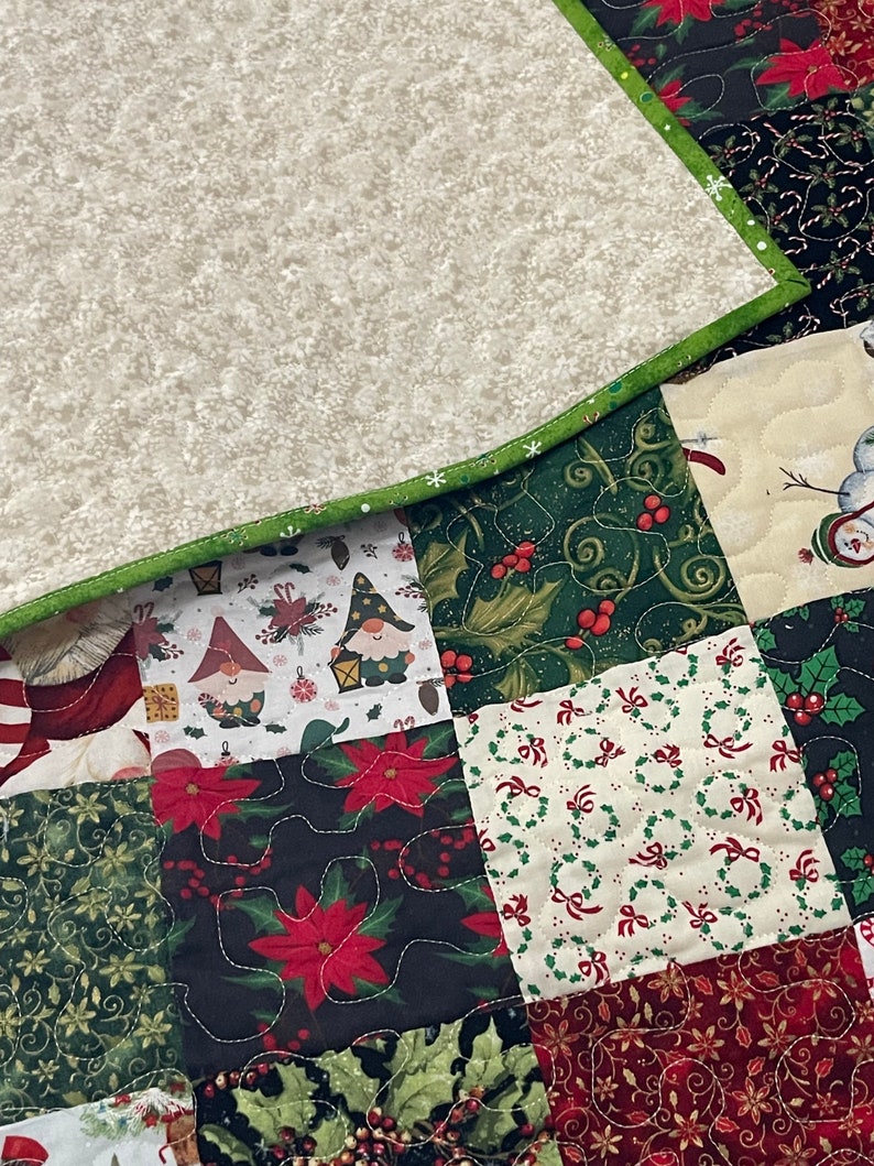 May include: A patchwork quilt with a variety of Christmas-themed patterns. The quilt features squares with gnomes, poinsettias, holly, and wreaths. The border is green, and the backing is a light beige with a snowflake pattern.