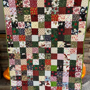 May include: A handmade Christmas quilt featuring a patchwork design. The quilt is composed of numerous squares in various festive patterns, including Santa Claus, poinsettias, and snowflakes. The color palette includes red, green, white, and black, with a green border.