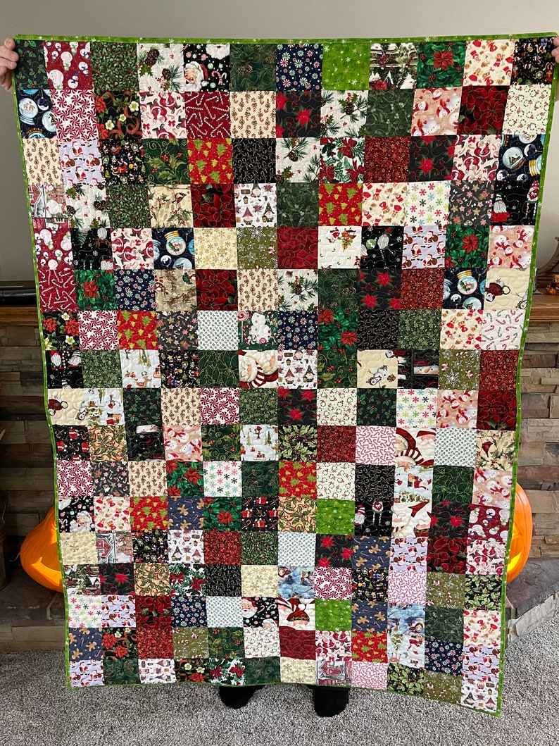 May include: A handmade Christmas quilt featuring a patchwork design. The quilt is composed of numerous squares in various festive patterns, including Santa Claus, poinsettias, and snowflakes. The color palette includes red, green, white, and black, with a green border.
