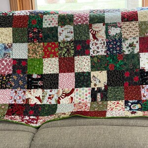 May include: A Christmas-themed patchwork quilt with a variety of festive patterns. The quilt features squares of red, green, white, and black fabrics, adorned with images of Santa Claus, snowmen, poinsettias, and candy canes. The quilt is edged with a green border.