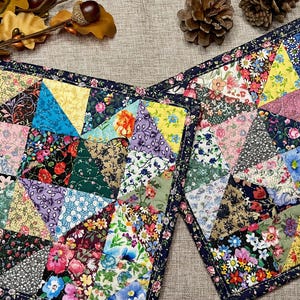 May include: Two square quilted potholders featuring a patchwork design of various floral and geometric fabric patterns in shades of blue, yellow, pink, green, and red. The potholders are finished with a dark blue fabric binding along the edges.