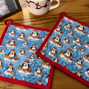 May include: Two red-trimmed blue quilted pot holders with a penguin and gift pattern.