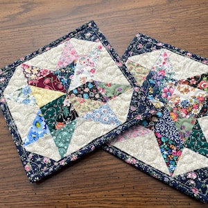 May include: Two handmade quilted hot pads. Each pad features a star design with a variety of floral fabric patches in shades of pink, blue, green, and yellow. The edges are finished with a dark blue floral border.