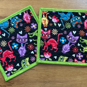 May include: Two square quilted fabric pot holders with a black background. The fabric is patterned with colorful cartoon cats, flowers, and other whimsical shapes. The edges are trimmed with a bright green binding.