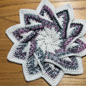 May include: A handmade, star-shaped crochet item in shades of white, gray, and pink. The crochet pattern features a spiral design with a scalloped edge. This decorative piece could be used as a trivet or coaster.