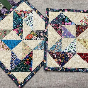 May include: Two square quilted potholders featuring a patchwork design of various floral and geometric fabric patterns in shades of red, blue, green, and yellow, set against a cream-colored background with a dark blue floral border trim.