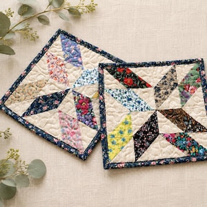 May include: Two square quilted fabric coasters featuring a star pattern made from various floral print patches in shades of blue, pink, yellow, red, and black, all bordered by a dark blue floral fabric binding with cream-colored quilted stitching.