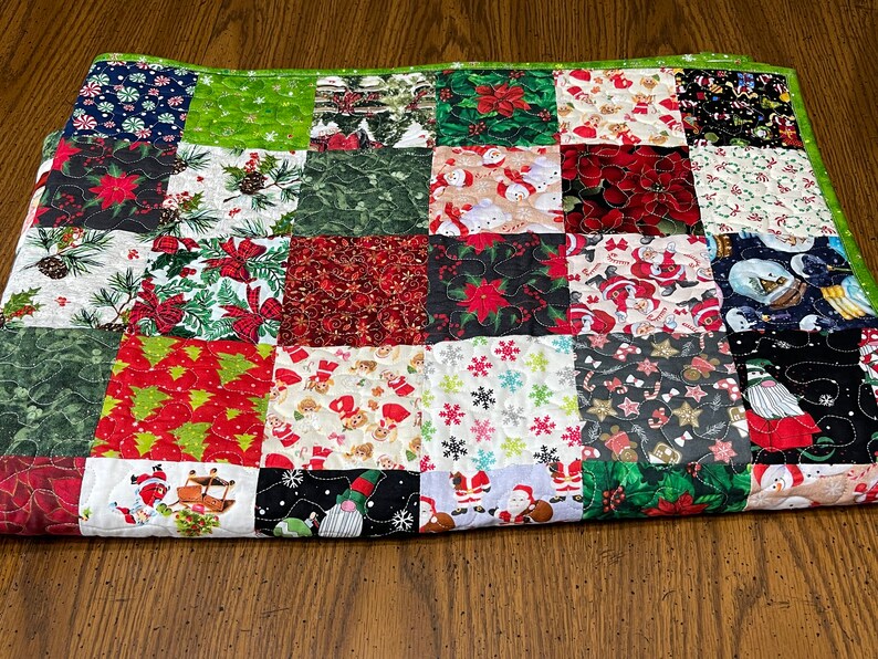 May include: A patchwork quilt with a green border, featuring various Christmas-themed fabrics. The squares display patterns of poinsettias, snowmen, Christmas trees, and Santa Claus, creating a festive and colorful design.