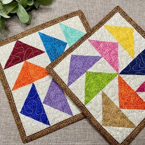 May include: Two square quilted fabric coasters featuring a patchwork design of multicolored triangles in shades of red, orange, blue, purple, yellow, pink, and green on a cream background, all bordered by a brown fabric trim with visible stitching.