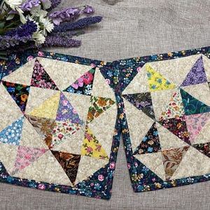 May include: Two square quilted potholders featuring a star pattern made of various floral fabric triangles in shades of pink, yellow, blue, green, and brown, set against a cream background with a dark blue floral border trim on all edges.