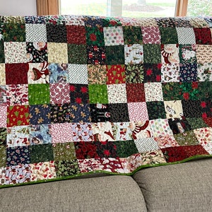 May include: A patchwork quilt with a green border, featuring a variety of Christmas-themed fabrics. The squares display patterns of Santa Claus, snowflakes, poinsettias, and candy canes. The quilt is draped over a neutral-colored sofa.