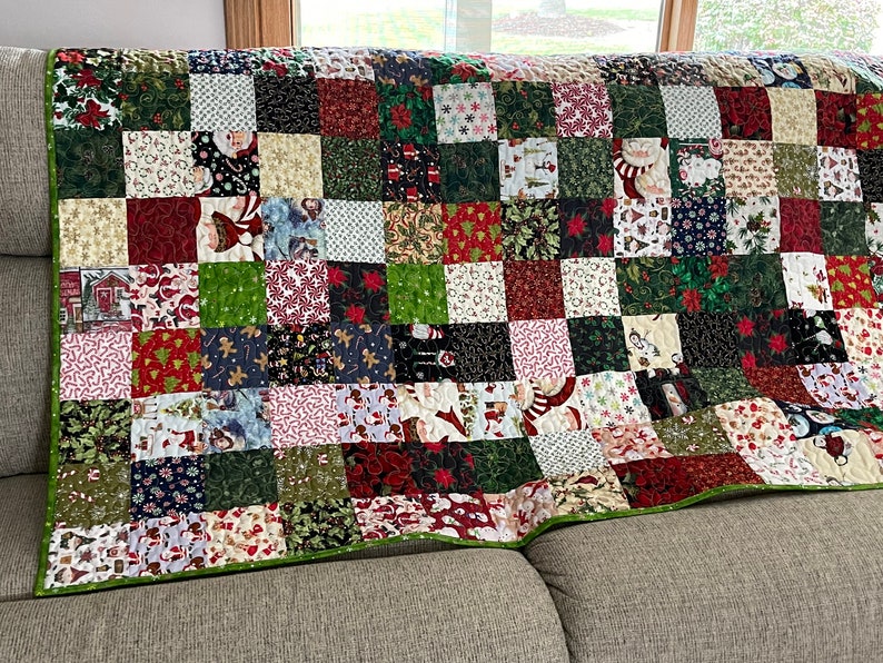 May include: A patchwork quilt with a green border, featuring a variety of Christmas-themed fabrics. The squares display patterns of Santa Claus, snowflakes, poinsettias, and candy canes. The quilt is draped over a neutral-colored sofa.