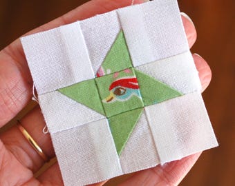 May include: A small, square quilt block featuring a green star design with a bird motif at the center. The star is surrounded by white fabric squares. The block is held in a hand, showcasing the detail of the patchwork.