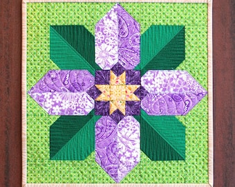 May include: A square quilted wall hanging featuring a stylized floral design. The central flower has purple petals with white floral patterns, green leaves, and a yellow starburst center. The background is a light green with a small dot pattern, and the piece is framed with a light-colored border.
