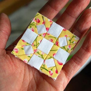 May include: A small, square patchwork quilt block held in a hand. The block features a yellow floral print fabric with white squares arranged in a grid pattern. The floral print includes pink flowers and green leaves.